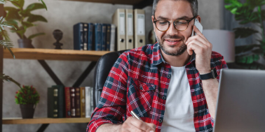 Smiling man writing notes while making phone call and using laptop at home