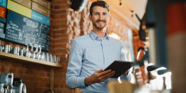 Handsome Caucasian Coffee Shop Owner is Working on Tablet Computer and Checking Inventory in a Cozy Loft-Style Cafe. Successful Restaurant Manager Standing Happy Behind Counter and Smiles on Camera.